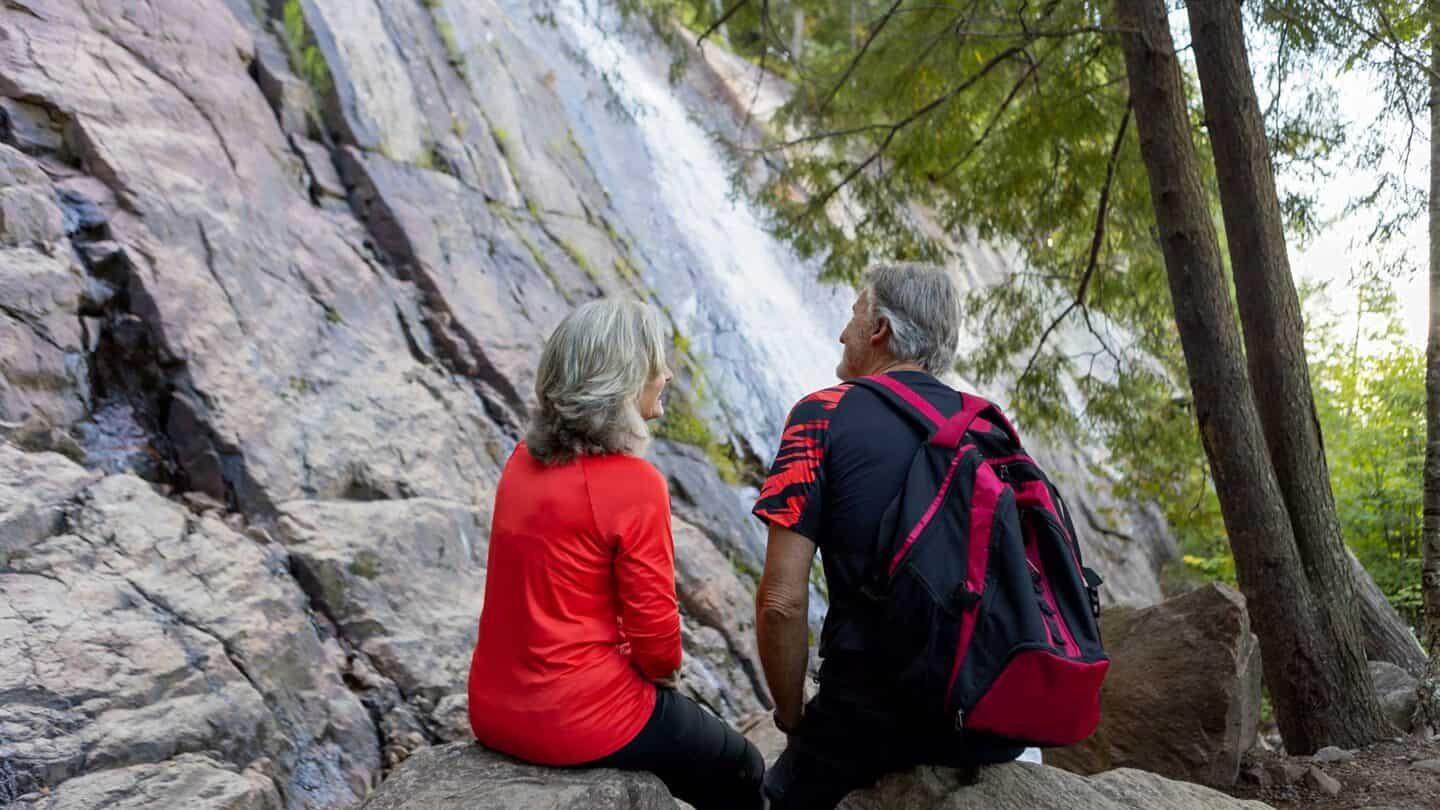 Randonnée en duo près d'une cascade en forêt, personnages assis sur une roche.