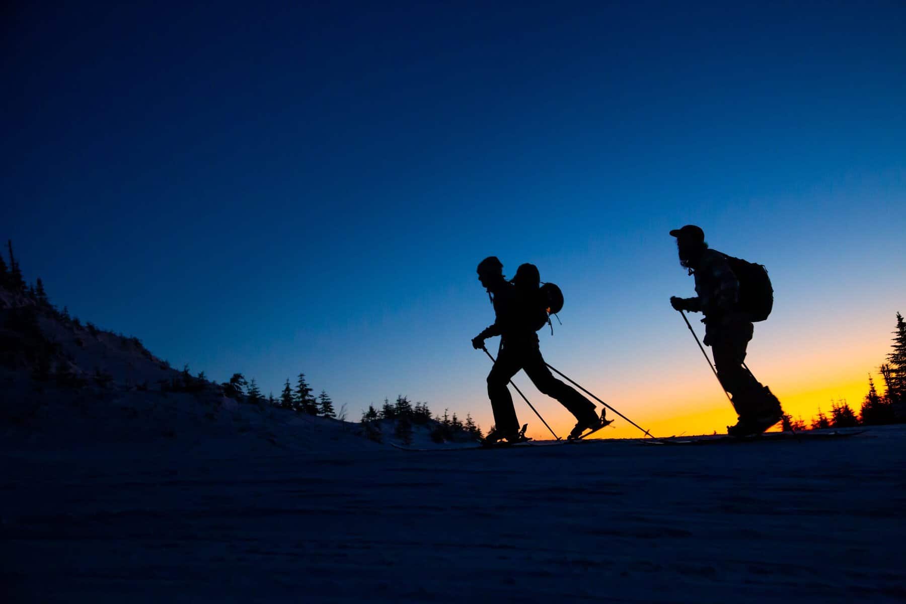 Two hikers skiing at dusk under a blue to orange gradient sky.