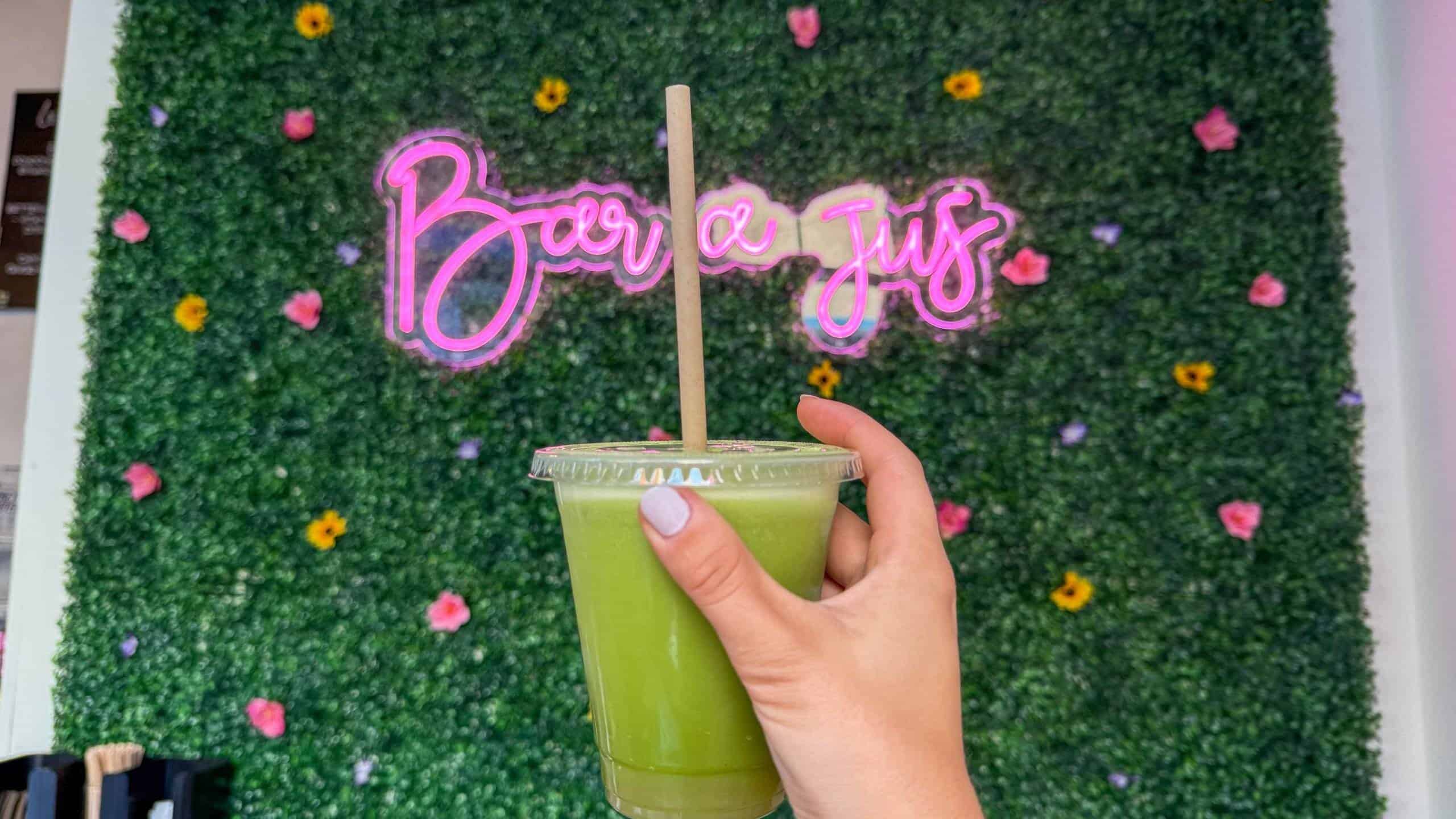 Hand holding a green juice with a pink neon sign in the background.