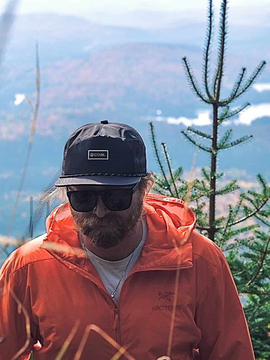 Homme portant veste orange et casquette sombre en forêt, ambiance montagne en arrière-plan.